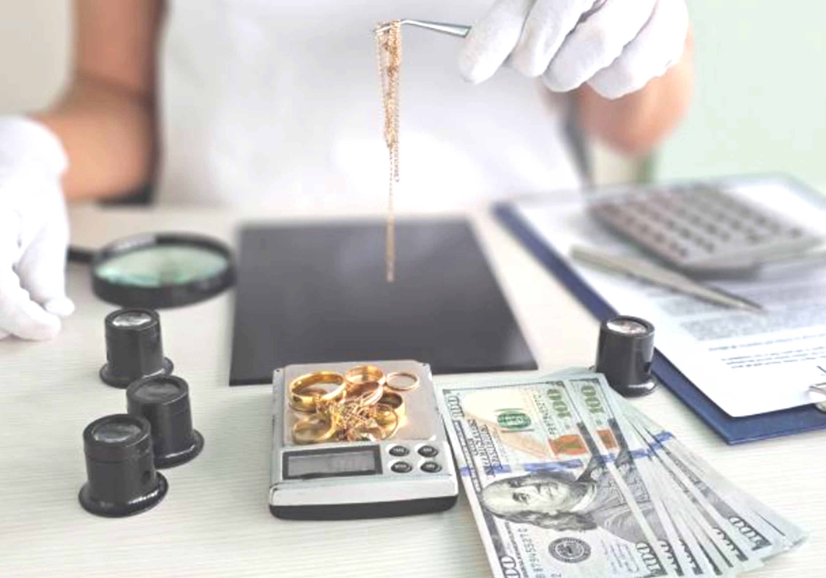 Person weighing gold jewelry on a scale with money and a magnifying glass nearby.