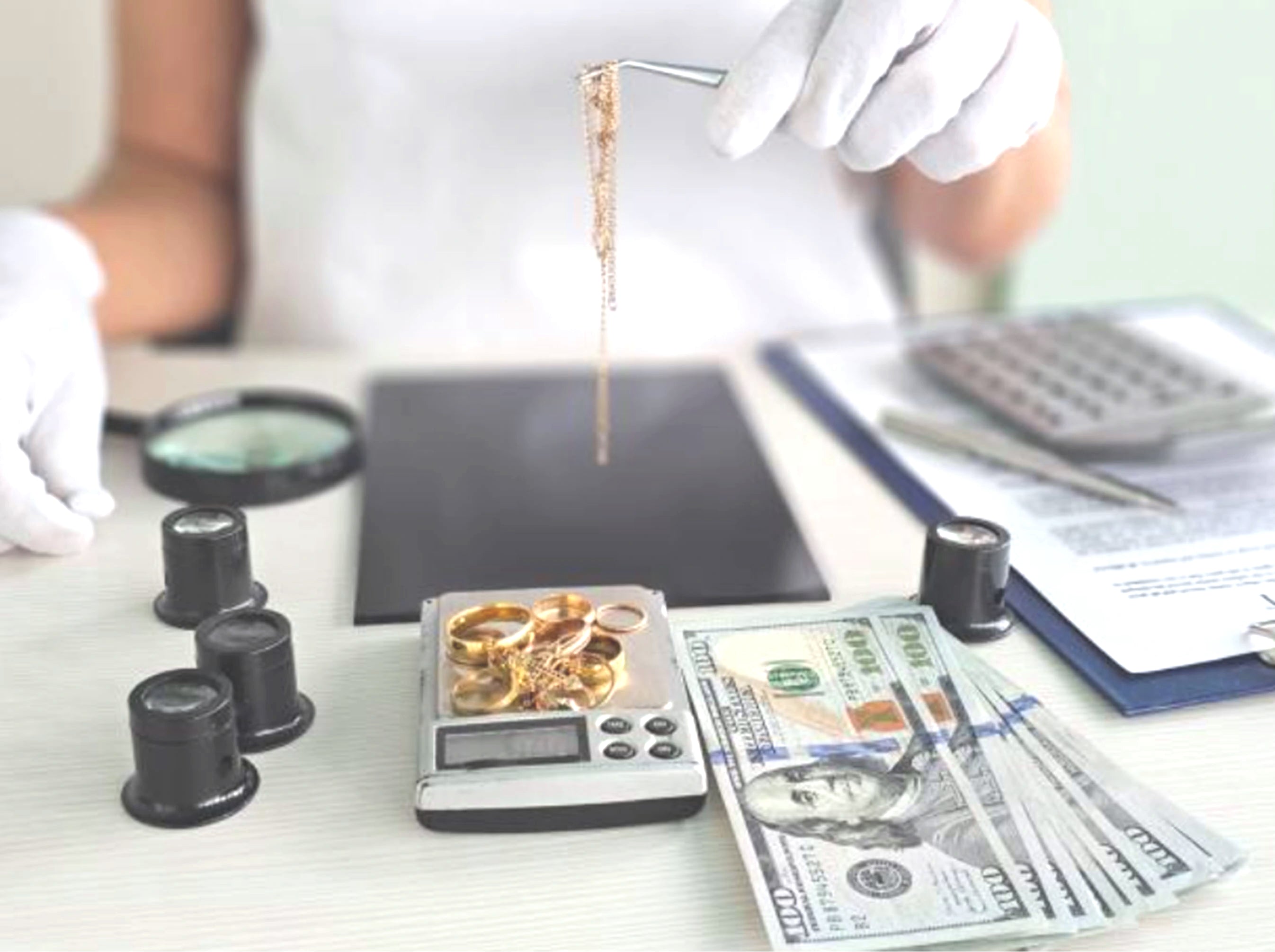 Person weighing gold jewelry on a scale with money and a magnifying glass nearby.
