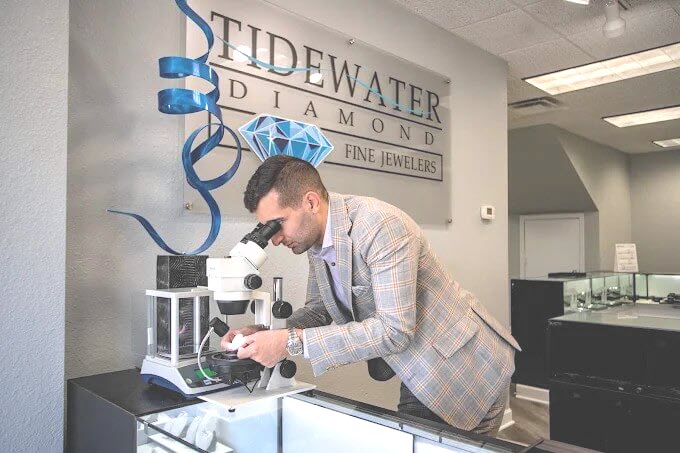 Man examining a diamond under a microscope in a jewelry store with 'Tidewater Diamonds' signage.